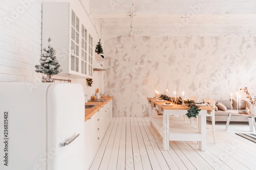 Loft style Apartment, large spacious living room with dining table and kitchen. Room with Christmas tree. Comfortable sofa, high large Windows. Light white brick wall.