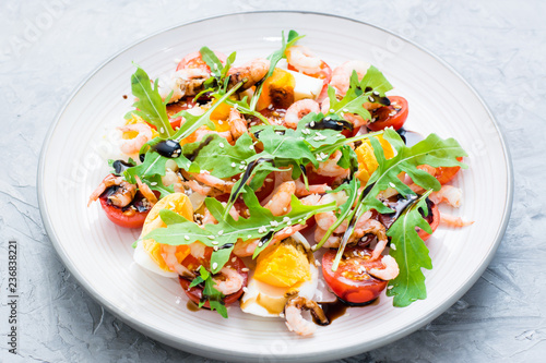 Ready-to-eat salad with cherry tomatoes, eggs, boiled shrimp, arugula, sesame seeds and balsamic sauce in a plate on the table