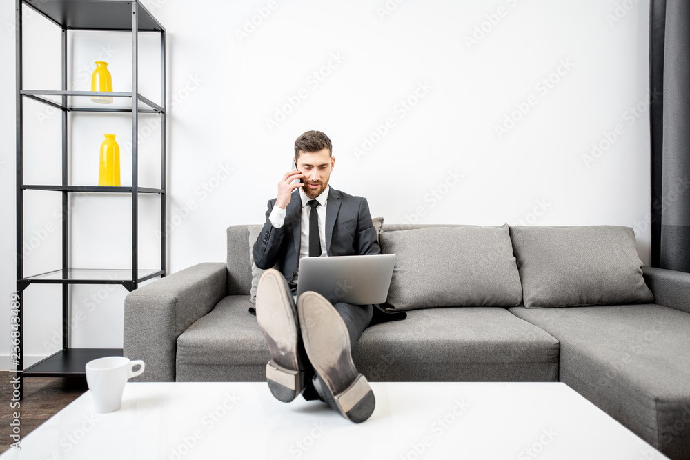 Elegant businessman dressed in the suit working with laptop and talking phone on the couch at home or comfortable office Elegant businessman dressed in the suit working with laptop and talking phone on the couch at home or comfortable office