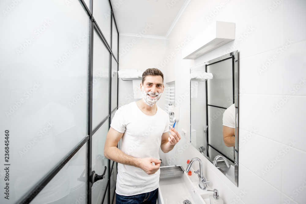 Portrait of a handsome man in white t-shirt shawing with blade and foam in the bathroom