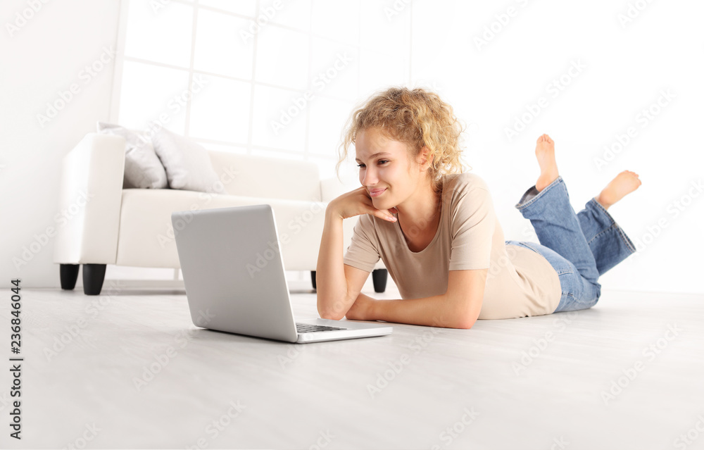 smiling beautiful young woman with computer laptop, lying on living room wooden floor in modern comfortable white home smiling beautiful young woman with computer laptop, lying on living room wooden floor in modern comfortable white home