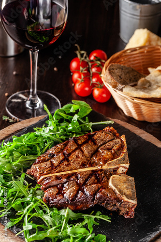 The concept of Italian cuisine. Juicy T-bone steak of beef aging stewed meat  with rucola salad and cherry tomatoes. A glass of red wine and bread basket on the table