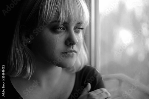Close up of crying depressed young blond woman near window at home. Sadness, nostlagic, depression. Black and white photo.