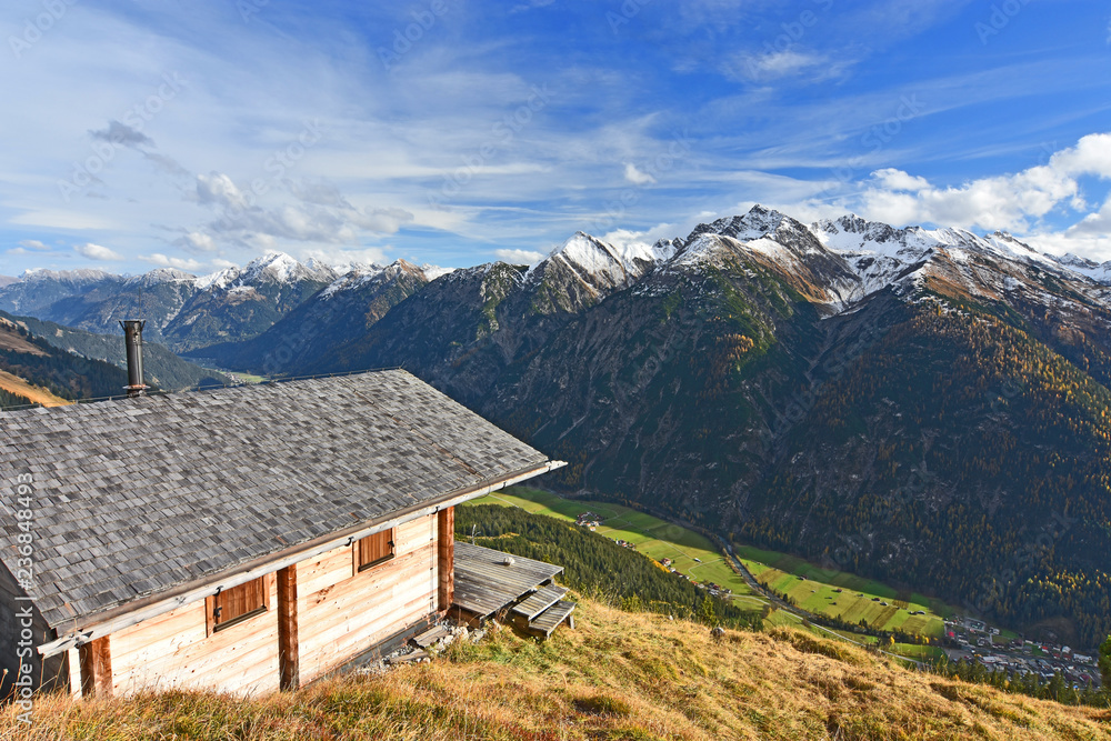 Wooden hut at a sunny day in autumn above the Lech valley with view to the Lechtal Alps, Tirol,Austria. Alpine landscape with green meadows, forest and snow-capped rocky mountains.