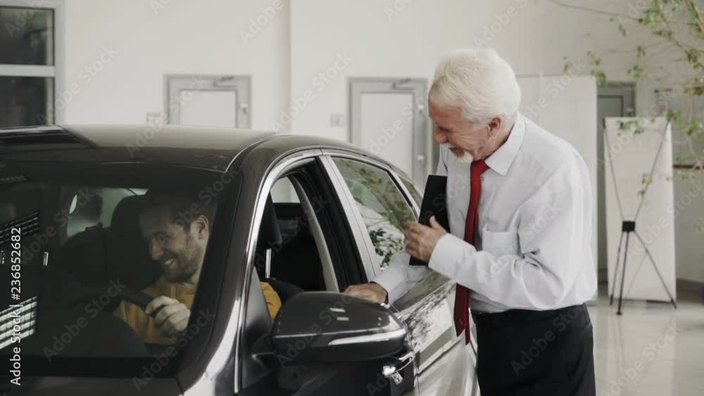 A Man get car keys from seller's hands. Happy customer is sitting in the car, just bought a car, in car dealership A Man get car keys from seller's hands. Happy customer is sitting in the car, just bought a car, in car dealership