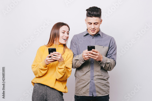 Young cheerful couple holding smartphones in their hands on white background. Girl looking in her boyfiend’s phones. photo