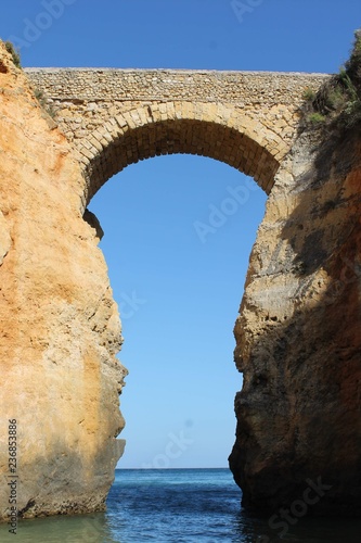 Beautiful view of an old stone bridge on a sunny beach with crystal clear blue water and fine white sand next to a steep rocky cliff in Lagos, Algarve, Portugal