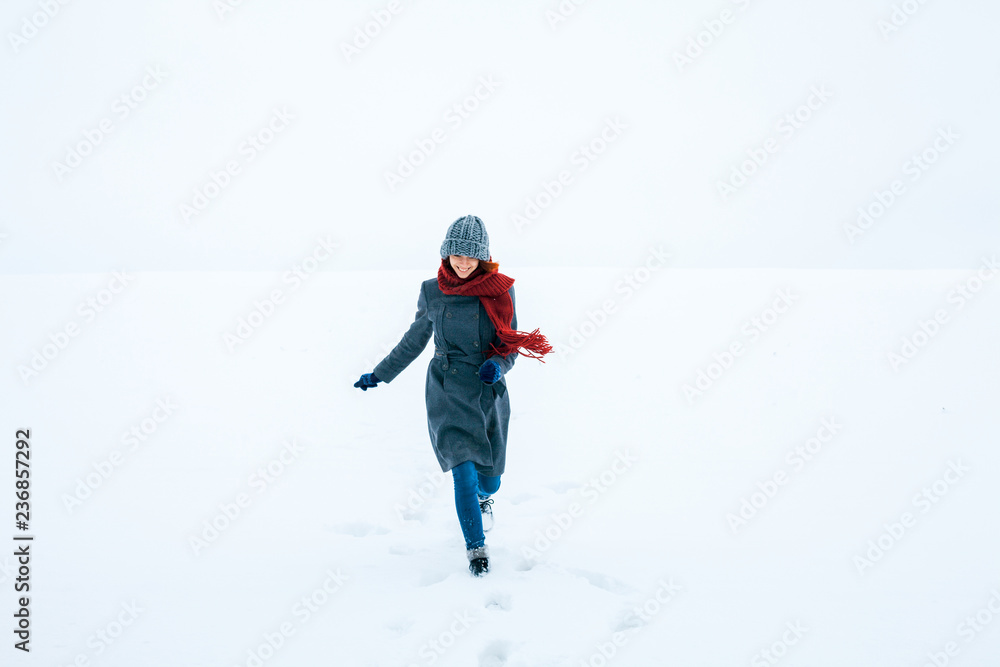 Happy smiling girl running in snow covered field. Model wearing winter coat, hat, red scarf. Copy, empty space for text