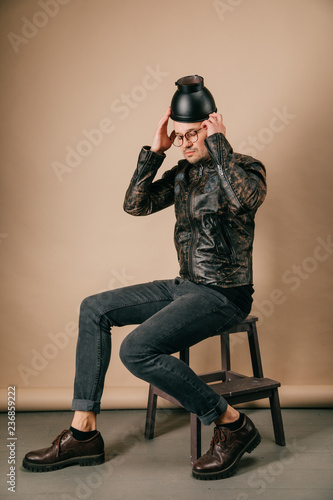 Funny portrait of young stylish man in fashionable clothes sitting on wooden chair with black pot on head in studio on brown background. Handsome smiling boy in glasses, leather jacket, brogue shoes.