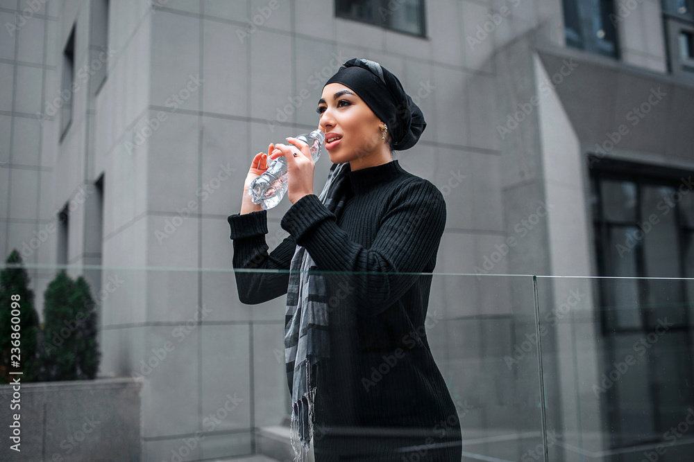 Fototapeta premium Attractive arabian woman drinks water from bottle. She looks straight forward. Woman stands at edge of glass railings.