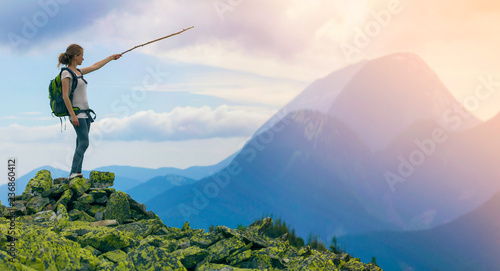 Young slim blond tourist girl with backpack points with stick at foggy mountain range panorama standing on rocky top on bright blue morning sky background. Tourism, traveling and climbing concept.