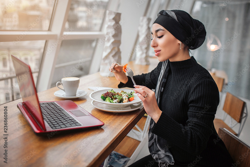 Nice young arabian woman sit at table in front of window. She eats salad. Laptop in on table.