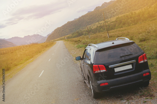 Black car SUV stands on the side of an asphalt road among the mountains. Travel by car through the mountain landscape. The rear of the vehicle with red lights in the foreground. © Yugan
