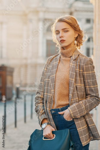 Outdoor fashion portrait of young beautiful fashionable girl wearing trendy checkered blazer, beige turtleneck, high-waisted jeans, wrist watch, carrying green blue handbag, posing in street 
