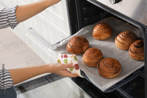 Beautiful young woman taking out tray of baked buns from oven in kitchen