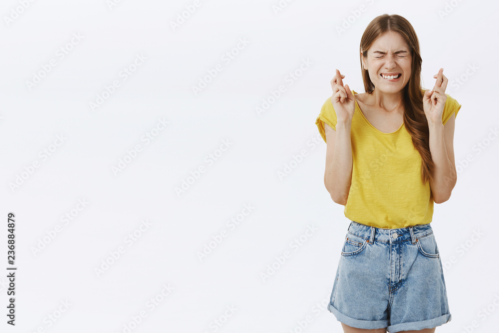 Portrait of charming worried european female student in yellow t-shirt and shorts clenching teeth closing eyes crossing fingers for good luck praying for wish come true having huge desire to win Portrait of charming worried european female student in yellow t-shirt and shorts clenching teeth closing eyes crossing fingers for good luck praying for wish come true having huge desire to win