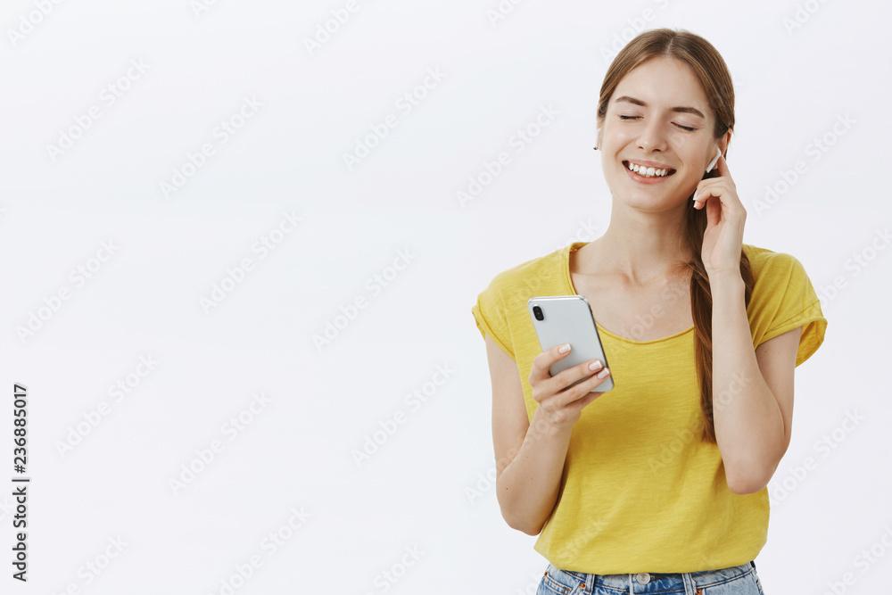 Delighted soft and tender charming female with braid holding smartphone wearing wireless earphones pressing earbud to ear while listening favorite song with closed eyes and delighted smile