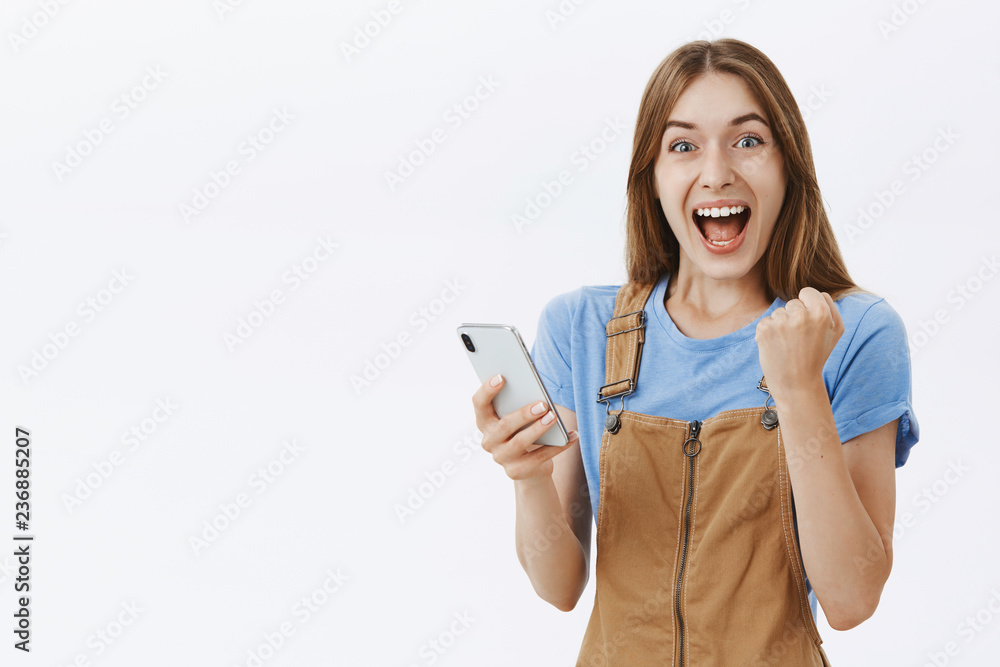 Waist-up shot of triumphing charming lucky girlfriend in brown overalls raising clenched fist in joy and victory celebrating new score in game holding smartphone hazing happy at camera over grey wall Waist-up shot of triumphing charming lucky girlfriend in brown overalls raising clenched fist in joy and victory celebrating new score in game holding smartphone hazing happy at camera over grey wall