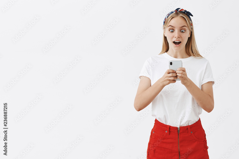 Stand still you gonna be shocked. Portrait of surprised and excited speechless girl with fair hair in skirt and headband popping eyes at smartphoen screen and gasping from amazement, reading Stand still you gonna be shocked. Portrait of surprised and excited speechless girl with fair hair in skirt and headband popping eyes at smartphoen screen and gasping from amazement, reading