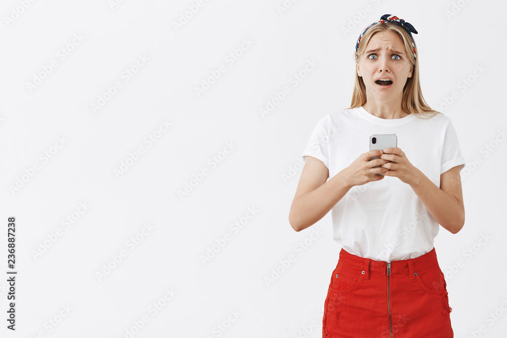 Studio shot of gloomy nervous and upset cute european female in red skirt frowning and grimacing from despair holding smartphone, reading shocking and disappointing news being worried over gray wall Studio shot of gloomy nervous and upset cute european female in red skirt frowning and grimacing from despair holding smartphone, reading shocking and disappointing news being worried over gray wall