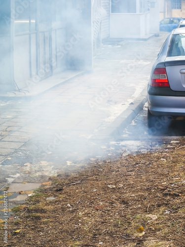 A large cloud of gray smoke from the exhaust pipe of a car in the courtyard of a residential building, ecology, urban smog, air pollution concept, vertical frame