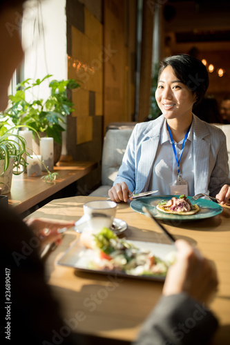 Smiling confident attractive young Asian businesswoman in stripped jacket sitting at table and smiling at colleague while having lunch with her in restaurant