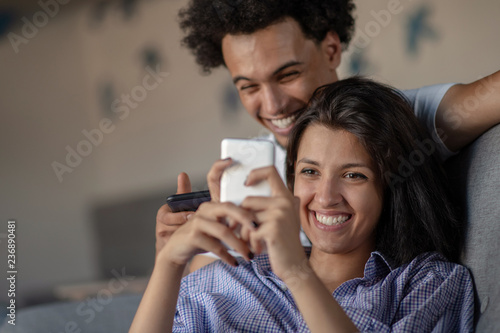 Attractive couple sitting on couch together looking at smartphone at home in the living room