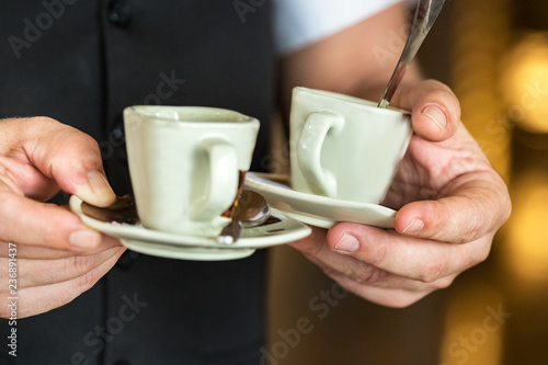 A waiter holding two cups of coffee with plate and spoon, indoors. photo