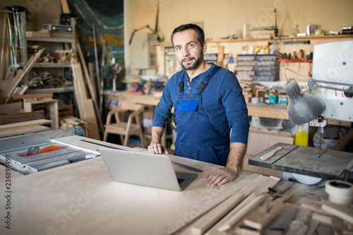 Waist up portrait of mature carpenter looking at camera and using laptop while working in joinery, copy space