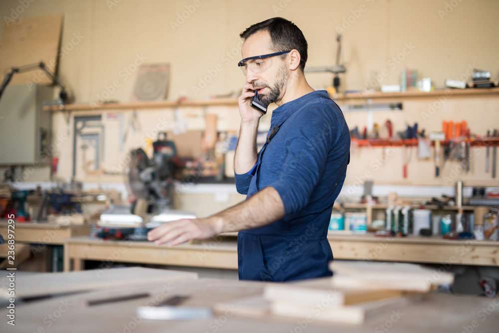 Side view portrait of mature craftsman speaking by phone while working in woodworking shop, copy space Side view portrait of mature craftsman speaking by phone while working in woodworking shop, copy space