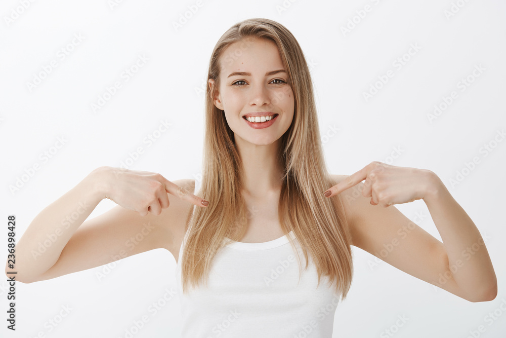 Waist-up shot of sincere and attractive friendly-looking pleasant woman with blond hair smiling broadly and pointing at herself as showing copy space on chest or downwards over gray background Waist-up shot of sincere and attractive friendly-looking pleasant woman with blond hair smiling broadly and pointing at herself as showing copy space on chest or downwards over gray background