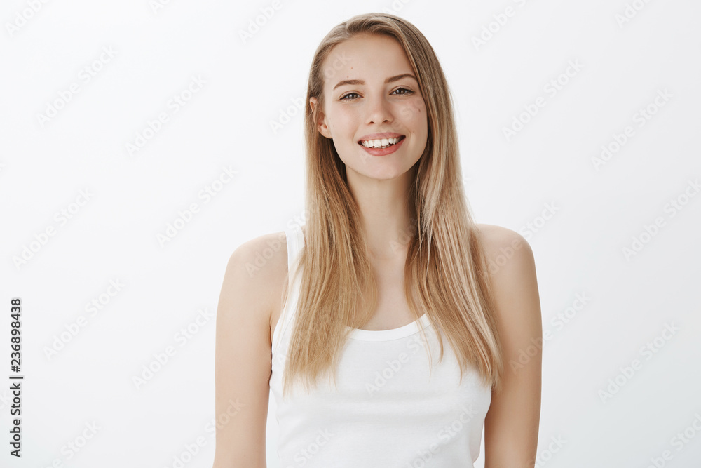 Waist-up shot of carefree joyful and attractive fair-haired european female student in white tank-top smiling delighted and happy looking ambitious at camera being in good mood over gray wall Waist-up shot of carefree joyful and attractive fair-haired european female student in white tank-top smiling delighted and happy looking ambitious at camera being in good mood over gray wall