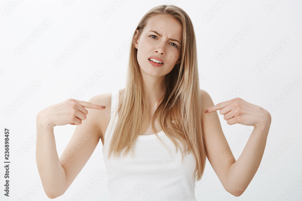 Waist-up shot of questioned and annoyed young attractive blonde in tank-top pointing at herself tilting head and frowning as being insulted with stupid pick-up line standing offended over gray wall Waist-up shot of questioned and annoyed young attractive blonde in tank-top pointing at herself tilting head and frowning as being insulted with stupid pick-up line standing offended over gray wall