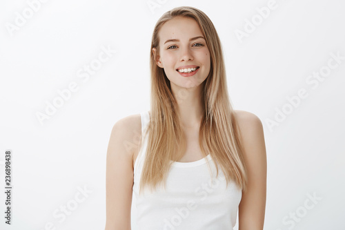 Waist-up shot of carefree joyful and attractive fair-haired european female student in white tank-top smiling delighted and happy looking ambitious at camera being in good mood over gray wall