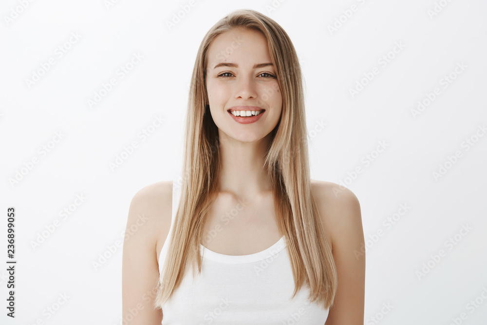 Waist-up shot of pleasant carefree and friendly attractive woman with fair hair and moles on clean pure face smiling broadly and gazing at camera with sincere happiness over gray background Waist-up shot of pleasant carefree and friendly attractive woman with fair hair and moles on clean pure face smiling broadly and gazing at camera with sincere happiness over gray background