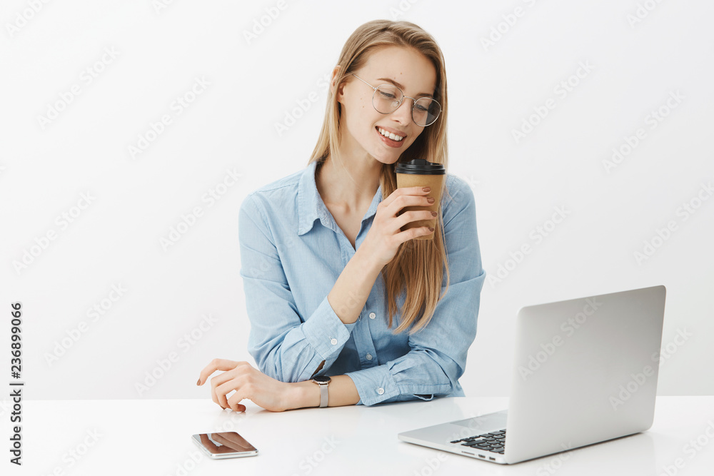 Girl addicted to work, smiling broadly as sipping coffee sitting near laptop and smartphone gazing at notebook screen, talking to friend via online video messages during office break over gray wall Girl addicted to work, smiling broadly as sipping coffee sitting near laptop and smartphone gazing at notebook screen, talking to friend via online video messages during office break over gray wall