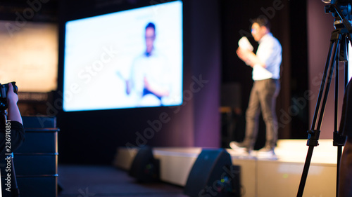 Presenter Presenting Presentation to Conference Audience. De-focused Blurred . Lecturer on Stage at Tech Forum. Speaker Giving Speech in Conference Hall Auditorium. Copy Space Screen Background.