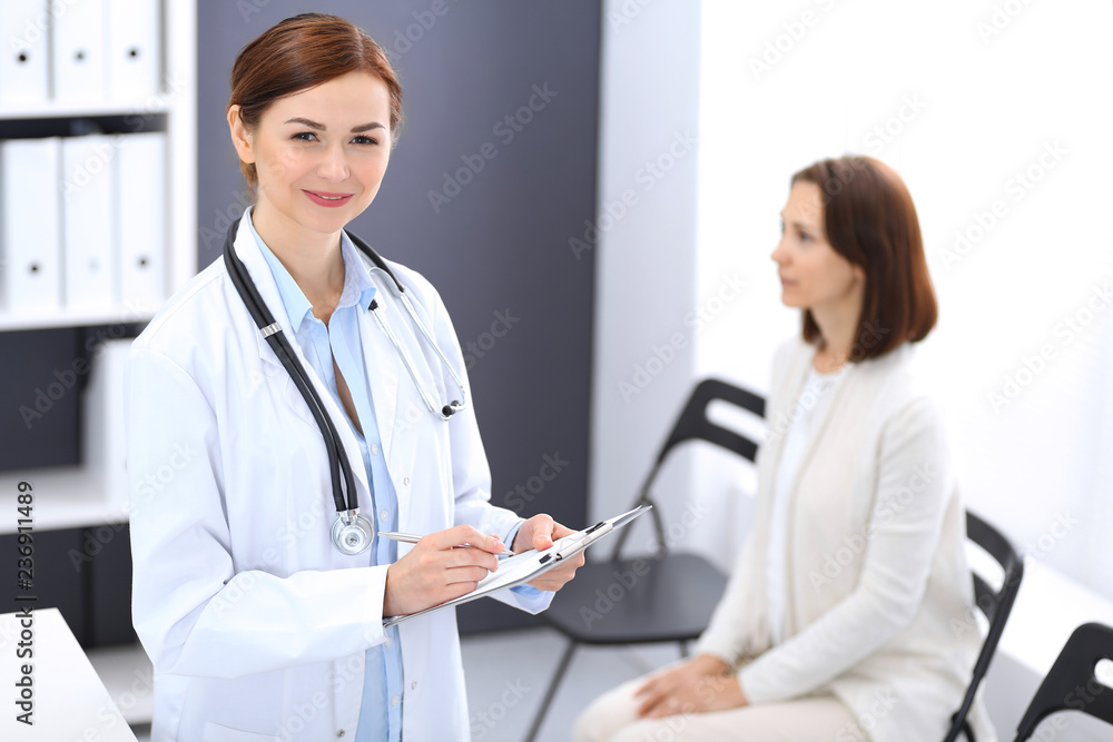 Doctor woman at work. Portrait of female physician filling up medical form while standing near reception desk at clinic or emergency hospital. Patient woman sitting at the background. Medicine concept Doctor woman at work. Portrait of female physician filling up medical form while standing near reception desk at clinic or emergency hospital. Patient woman sitting at the background. Medicine concept