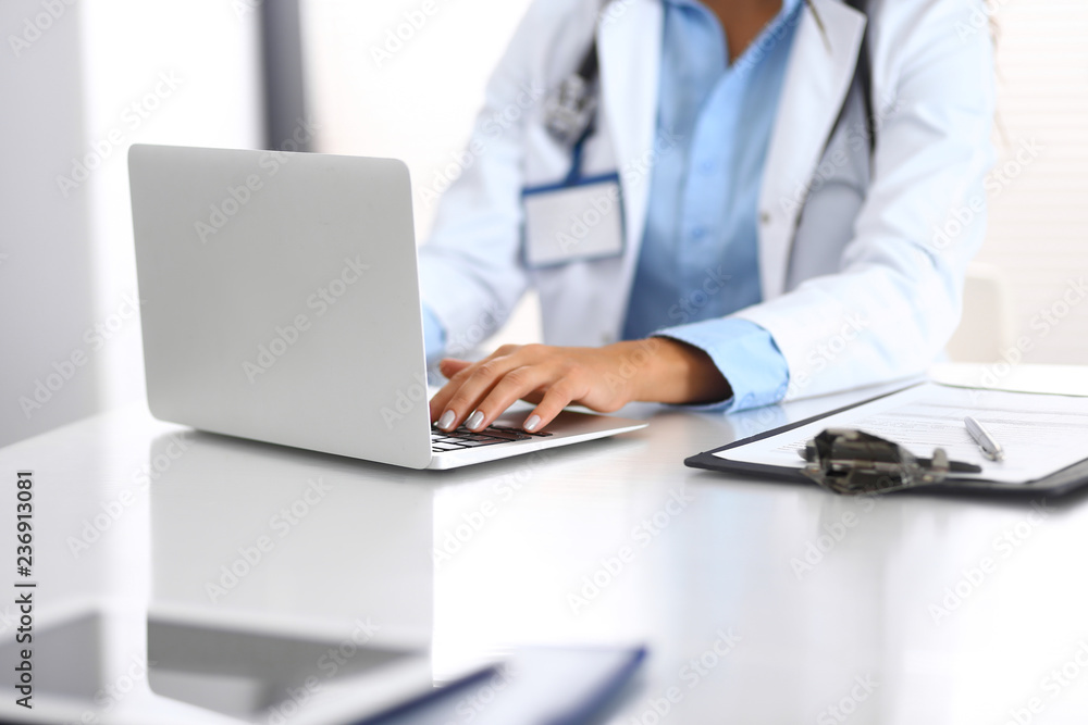 Fototapeta premium Unknown doctor woman typing on laptop computer while sitting at the desk in hospital office. close-up of hands. Physician at work. Medicine and health care concept