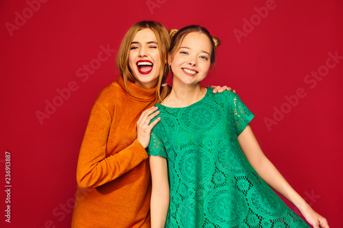 Two young beautiful blond smiling hipster girls posing in trendy summer clothes. Carefree women isolated on red background. Positive models going crazy and hugging