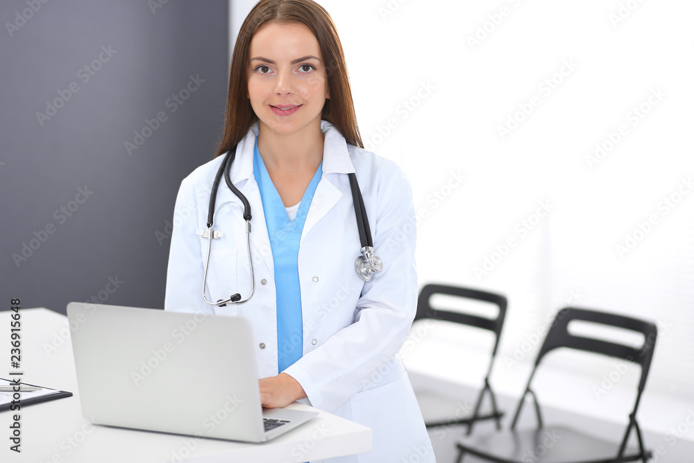 Doctor woman at work. Portrait of female physician using laptop computer while standing near reception desk at clinic or emergency hospital. Medicine and healthcare concept Doctor woman at work. Portrait of female physician using laptop computer while standing near reception desk at clinic or emergency hospital. Medicine and healthcare concept