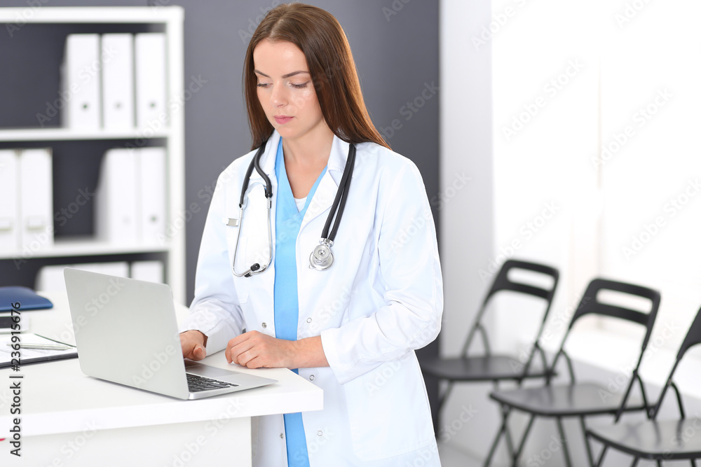 Doctor woman at work. Portrait of female physician using laptop computer while standing near reception desk at clinic or emergency hospital. Medicine and healthcare concept Doctor woman at work. Portrait of female physician using laptop computer while standing near reception desk at clinic or emergency hospital. Medicine and healthcare concept