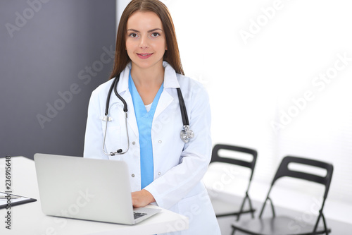 Doctor woman at work. Portrait of female physician using laptop computer while standing near reception desk at clinic or emergency hospital. Medicine and healthcare concept