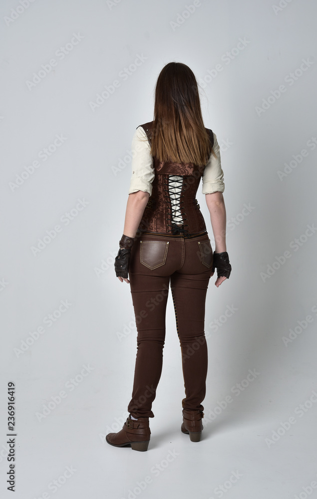 full length portrait of brunette girl wearing brown leather steampunk outfit. standing pose with back to the camera, on grey studio background. full length portrait of brunette girl wearing brown leather steampunk outfit. standing pose with back to the camera, on grey studio background.