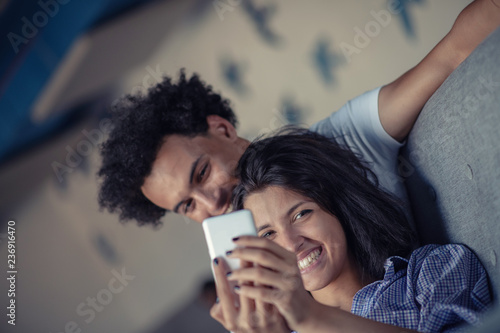 Attractive couple sitting on couch together looking at smartphone at home in the living room