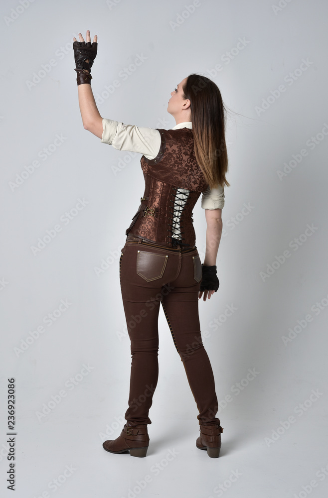 full length portrait of brunette  girl wearing brown leather steampunk outfit. standing pose with back to the camera, on grey studio background.