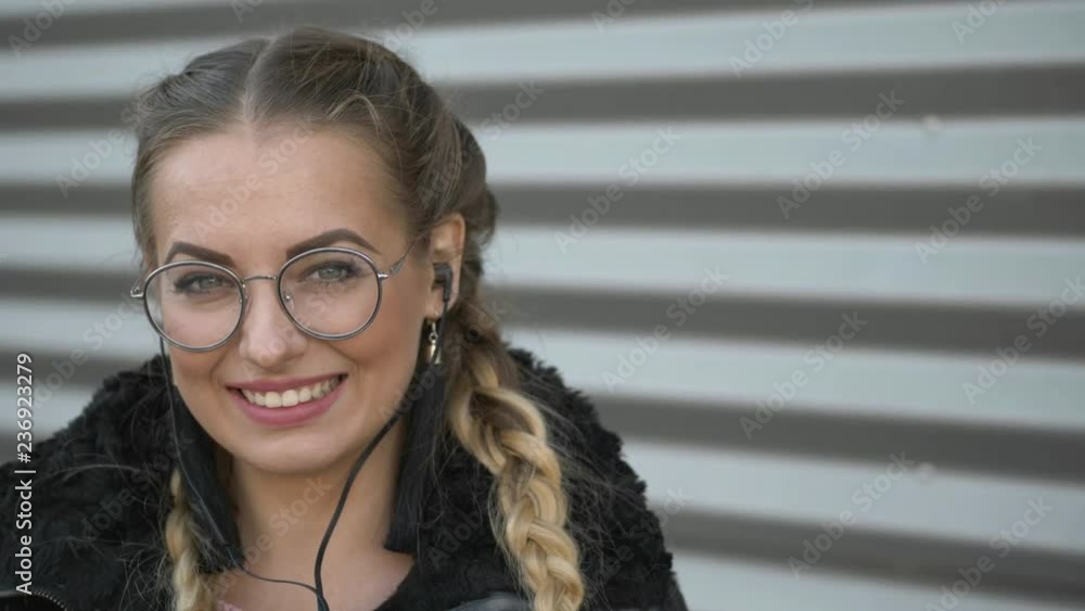 Slow motion portrait of beautiful young blonde woman in glasses, braids hairstyle, in smartphone headphones enjoying music outdoors. Posing against urban metal strips background. Slow motion portrait of beautiful young blonde woman in glasses, braids hairstyle, in smartphone headphones enjoying music outdoors. Posing against urban metal strips background.