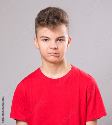 Emotional portrait of caucasian upset problem teen boy. Sad boy looking at camera. Worried  child wearing red t-shirt, on gray background.