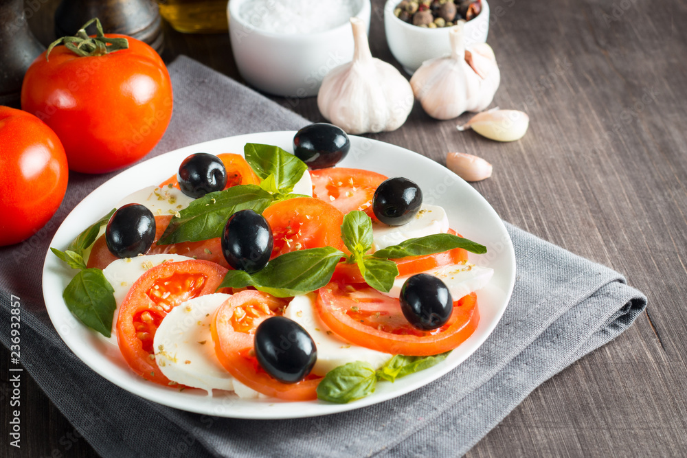 Photo of Caprese Salad with tomatoes, basil, mozzarella, olives and olive oil on wooden background. Italian traditional caprese salad ingredients. Mediterranean, organic and natural food concept.