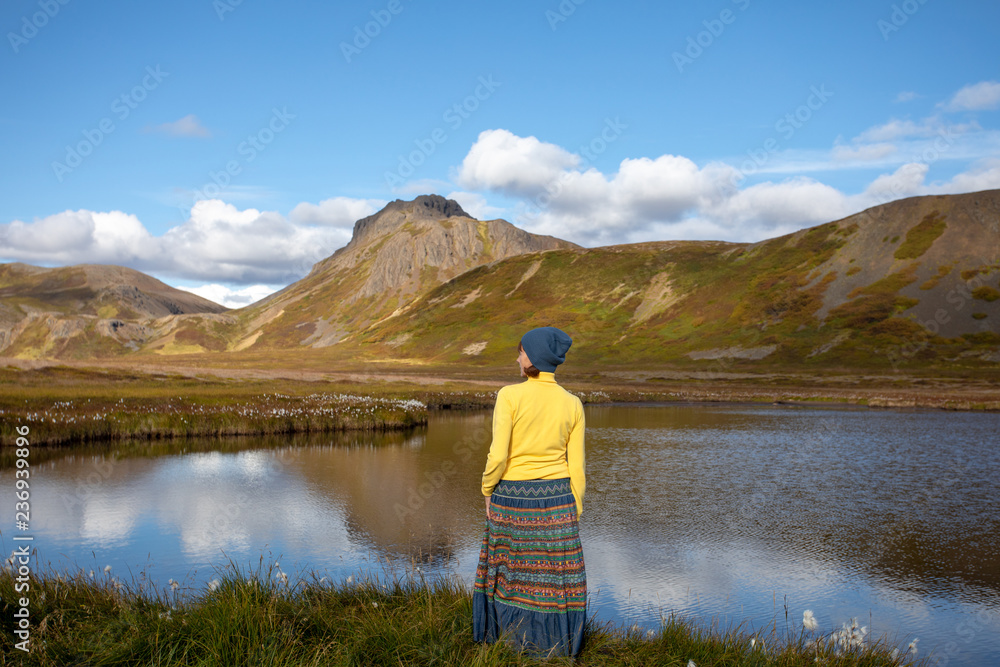 A young woman admires the view on the shore of a mountain lake. Clouds of mountains, and blue sky reflected in water. Summer view of travel. Iceland Europe